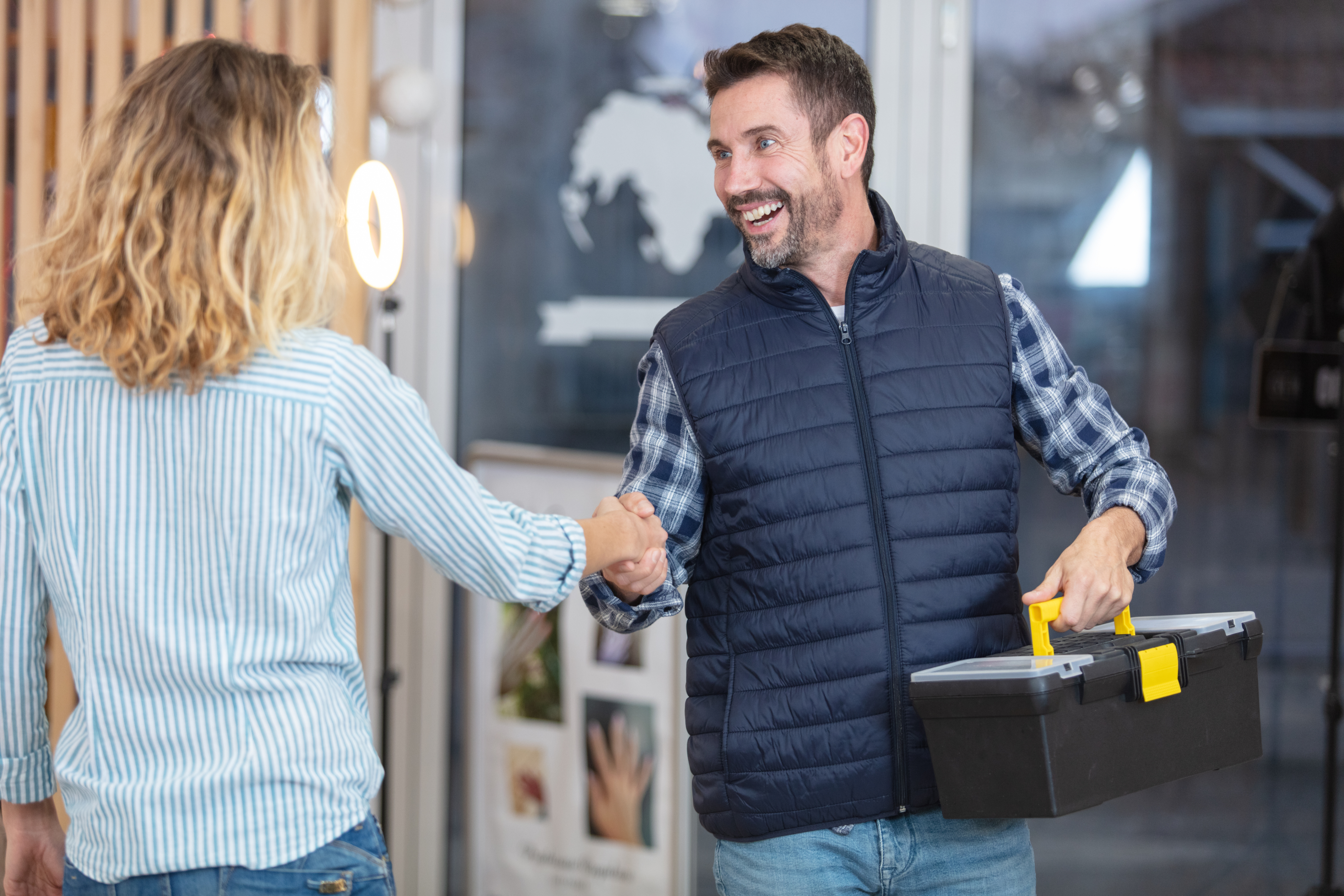 A smiling man in a navy vest, holding a toolbox, shakes hands with a woman in a striped shirt. 